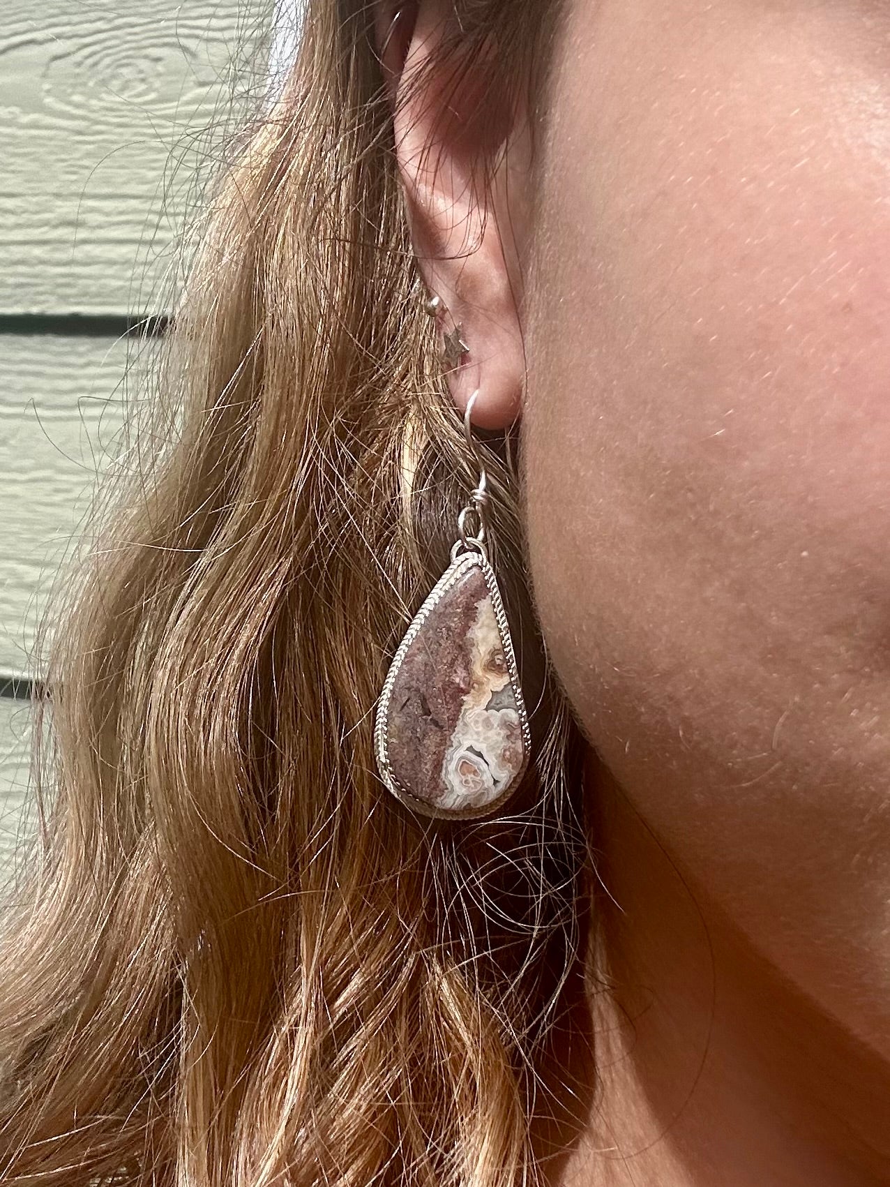 A close-up of a person wearing a teardrop-shaped dangle earring with a natural agate stone bezel set in silver